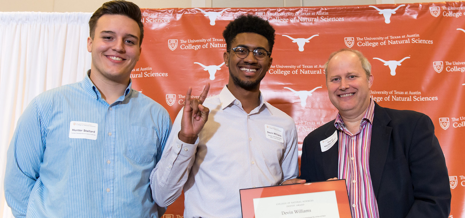 student shows hook-em-horns sign with instructor and dean as receiving an award in front of Longhorn backdrop